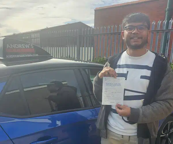 Anthony stands next to a blue learner car outside rew’s Driving School signage visible on the roof.