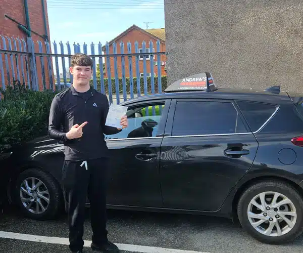 Kegan stands beside a black driving school car at ar a fenced area and buildings in the background.