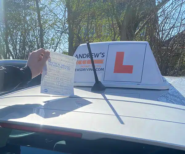 Pass certificate held up beside an Andrew’s Drivinate at Wrexham Driving Test Centre on a sunny day