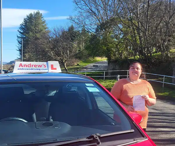 MJ holding her pass certificate beside a red learnmatic driving test in Rhyl on a bright sunny day.