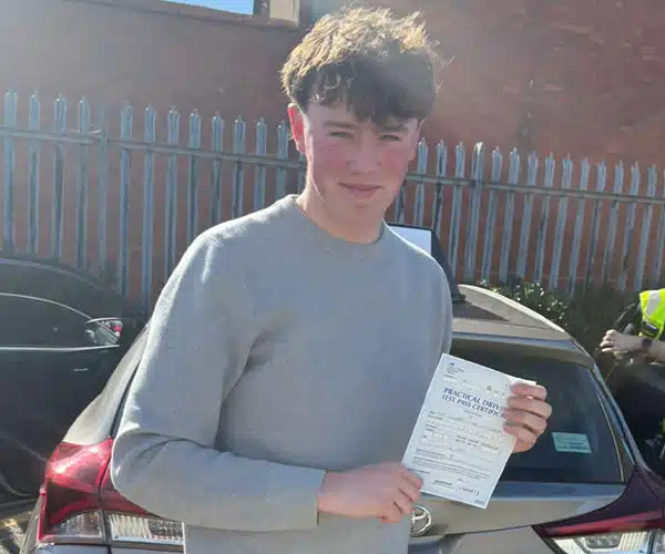Teddy holding his driving test pass certificate be outside Rhyl Driving Test Centre on a sunny day.