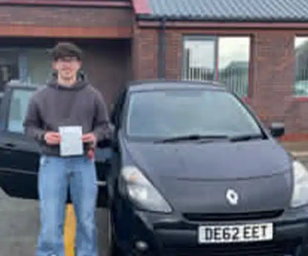 Alfie holding driving test pass certificate outside Rhyl Driving Test Centre next to black Renault