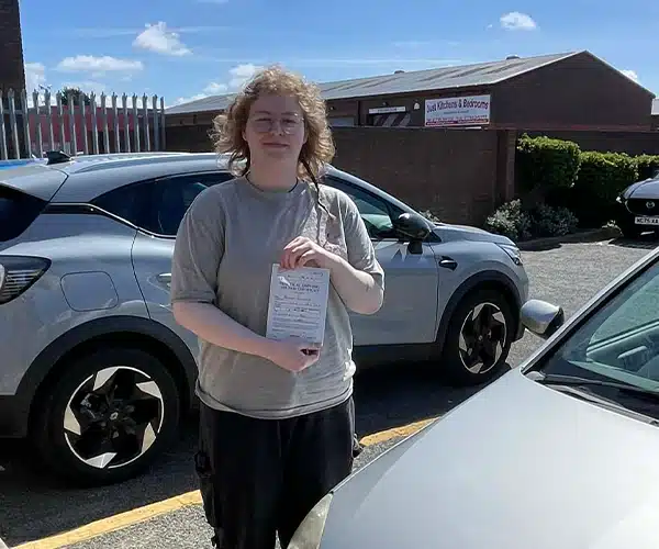 Briana holding her driving test pass certificate at Rhyl Driving Test Centre next to a learner car