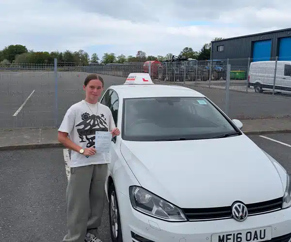 Maisy holding her driving test pass certificate ator Driving Test Centre beside a white learner car
