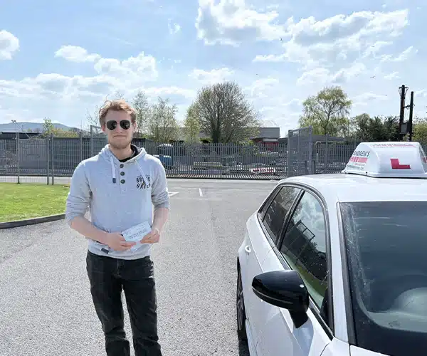 Richard from Llandudno holding his pass certificatt Bangor Driving Test Centre beside a learner car