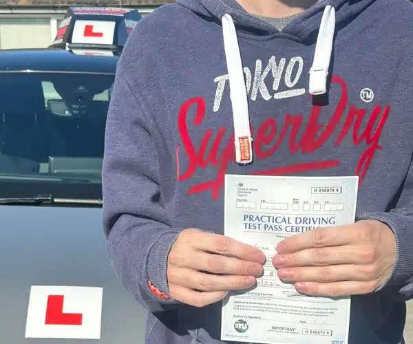 Sean holding practical driving test pass certificate at Rhyl Driving Test Centre with learner car